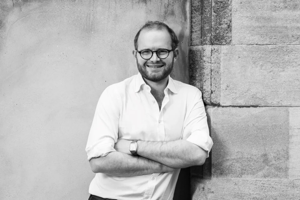 Portrait image of Dr Oliver Cox in front of a wall