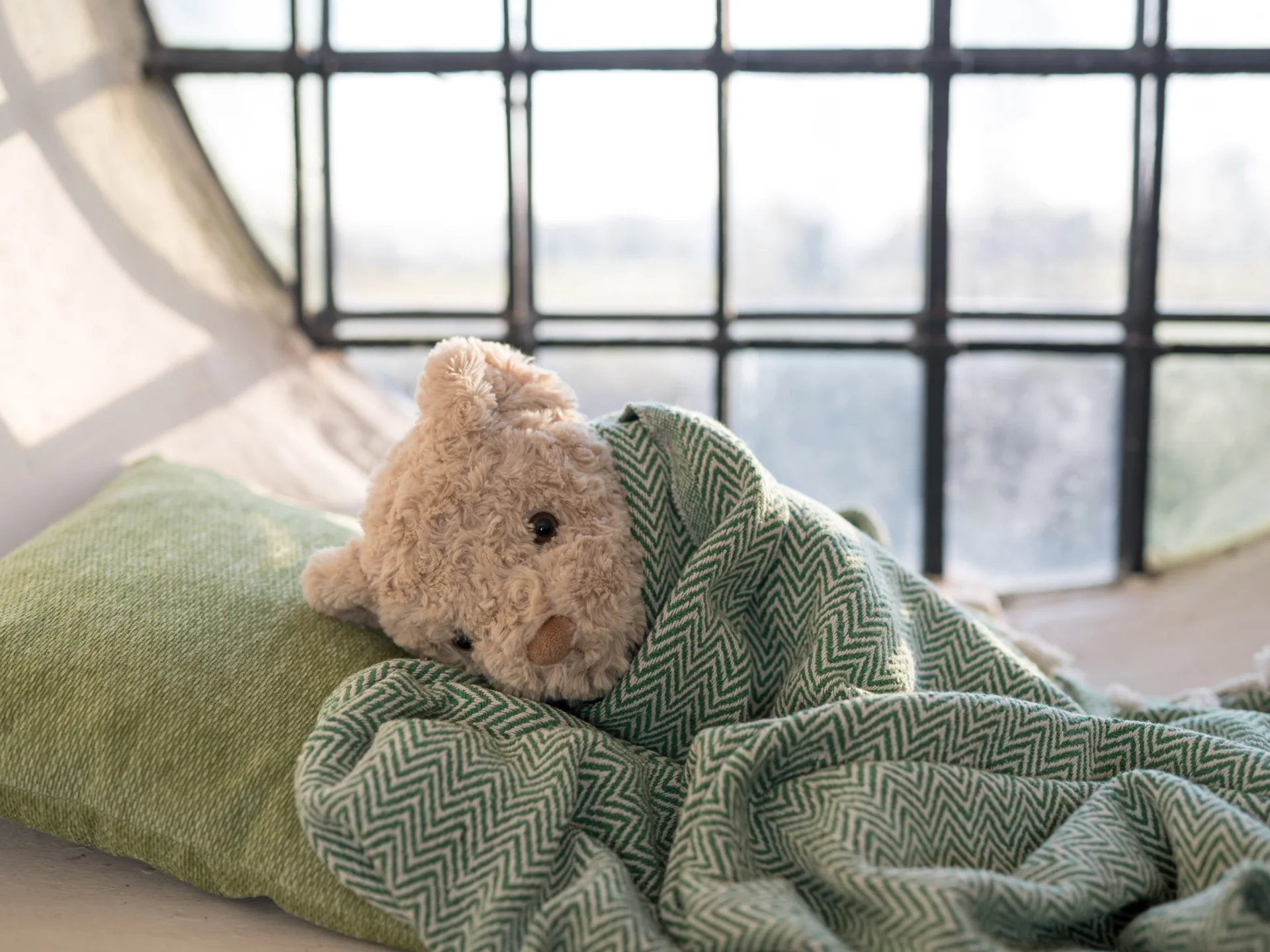 Cosy teddybear lying on pillow wrapped up in blanket in front of a clear glass circular window