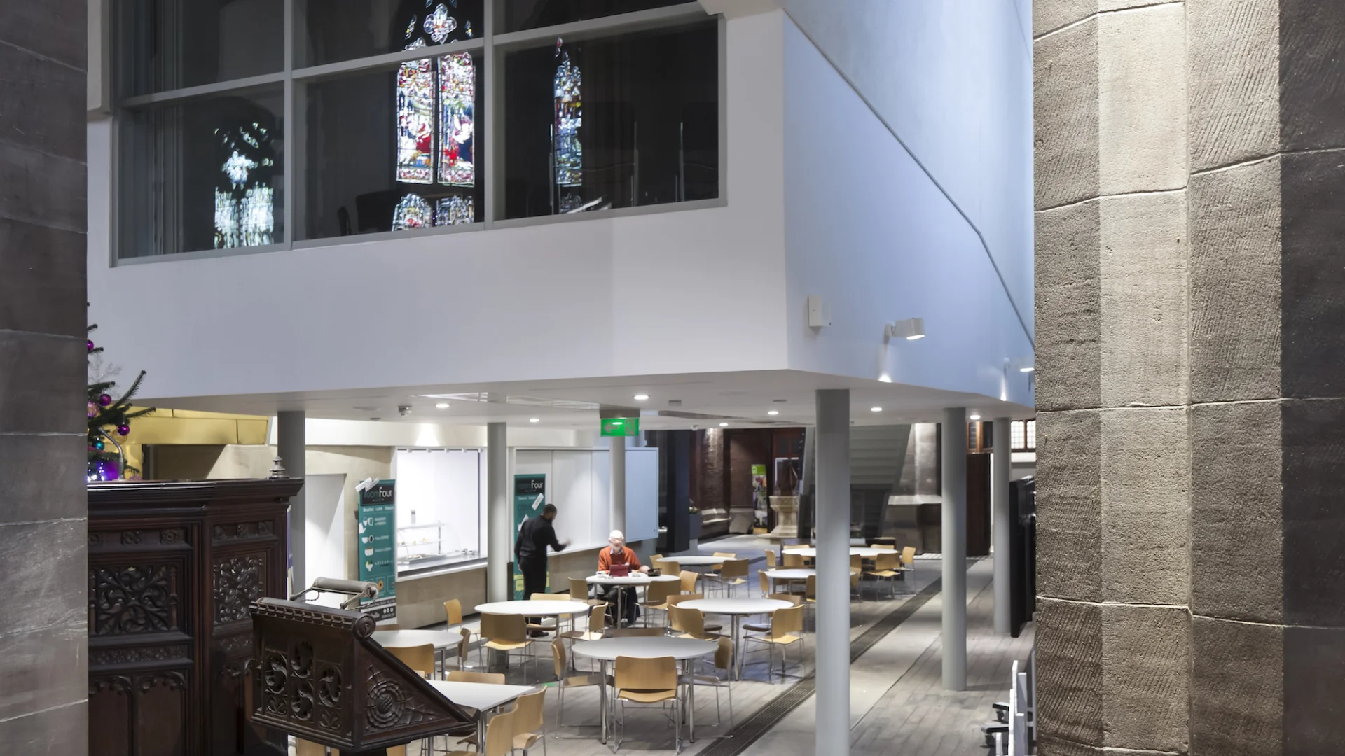 Shows tables and a canteen with people eating in the 'building within a building'. Through this you can see the original stained glass.