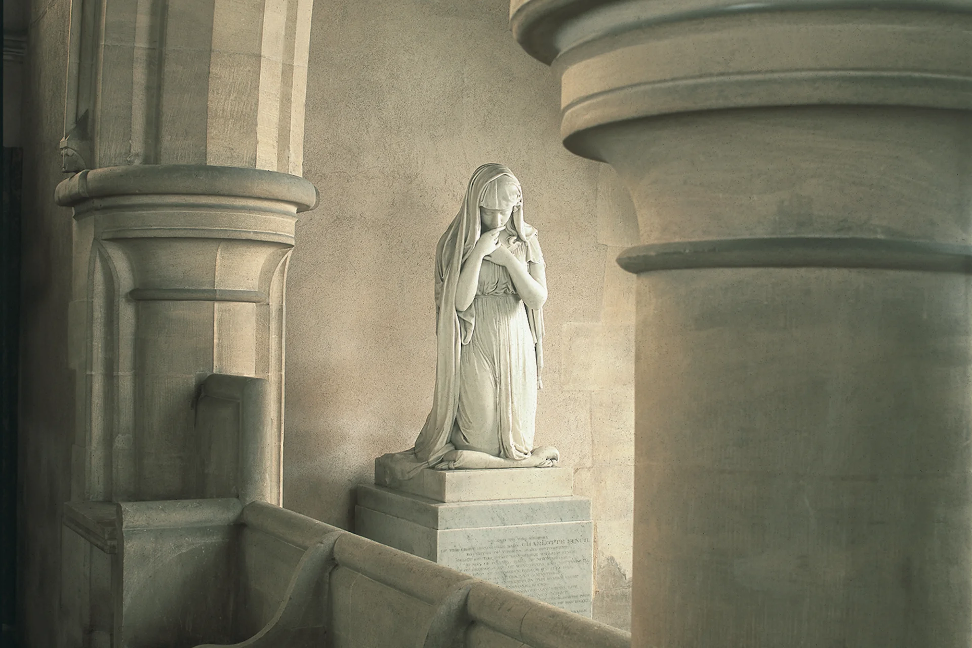Beautiful marble statue of a woman with her hands to her lips and chest with veil pulled back. The statue is at the side of the church between the arches.