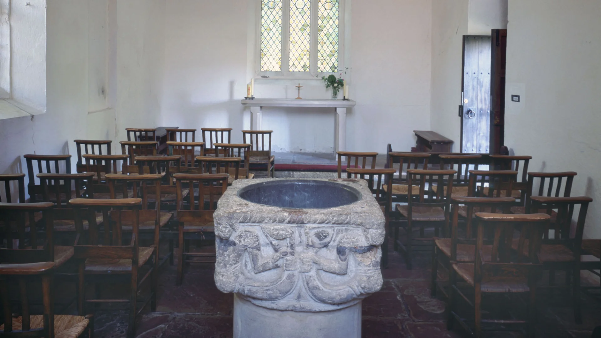 Stone font engraved with two mermen in a white washed church interior with small window above simple altar and wooden chairs.