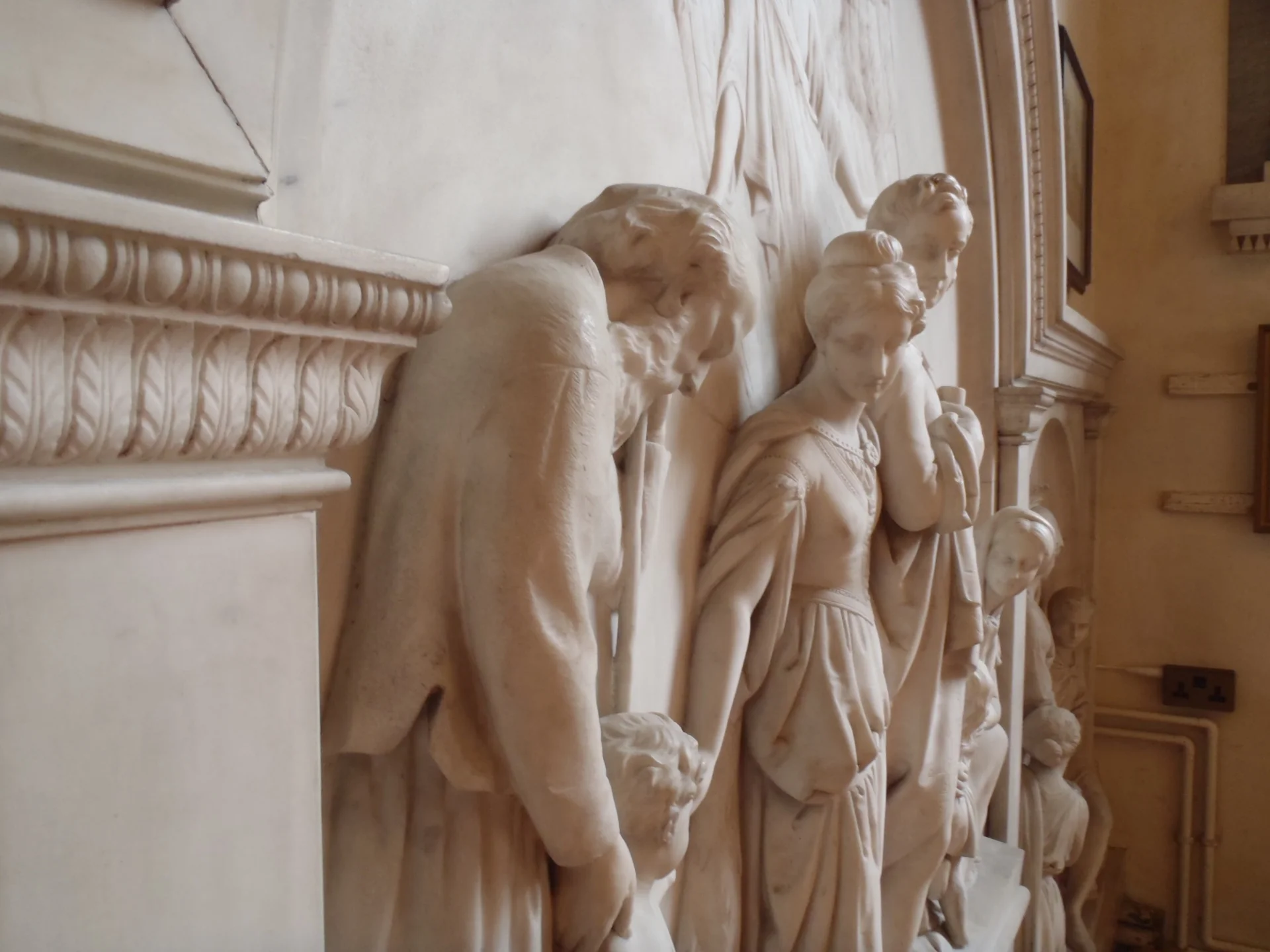White marble wall monument. Image of  figures taken from the side so that the woman's face is visible and behind the back of a man's head, you see the faces of children and her partner.