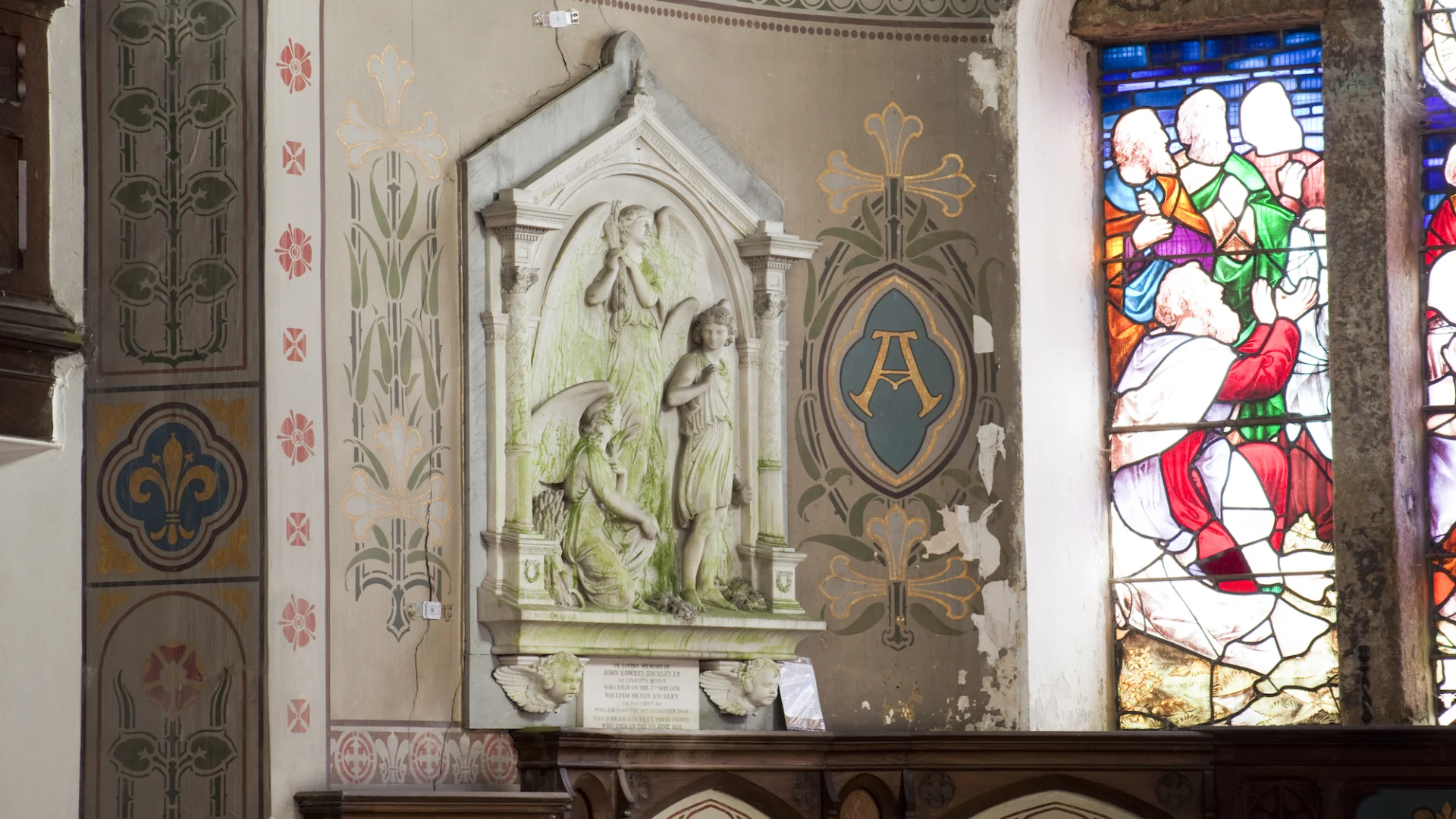 Ornate Wall Monument with angel. Wall Paintings blue with gold florals and red flowers. Edge of stained glass window visible depicting disciples.