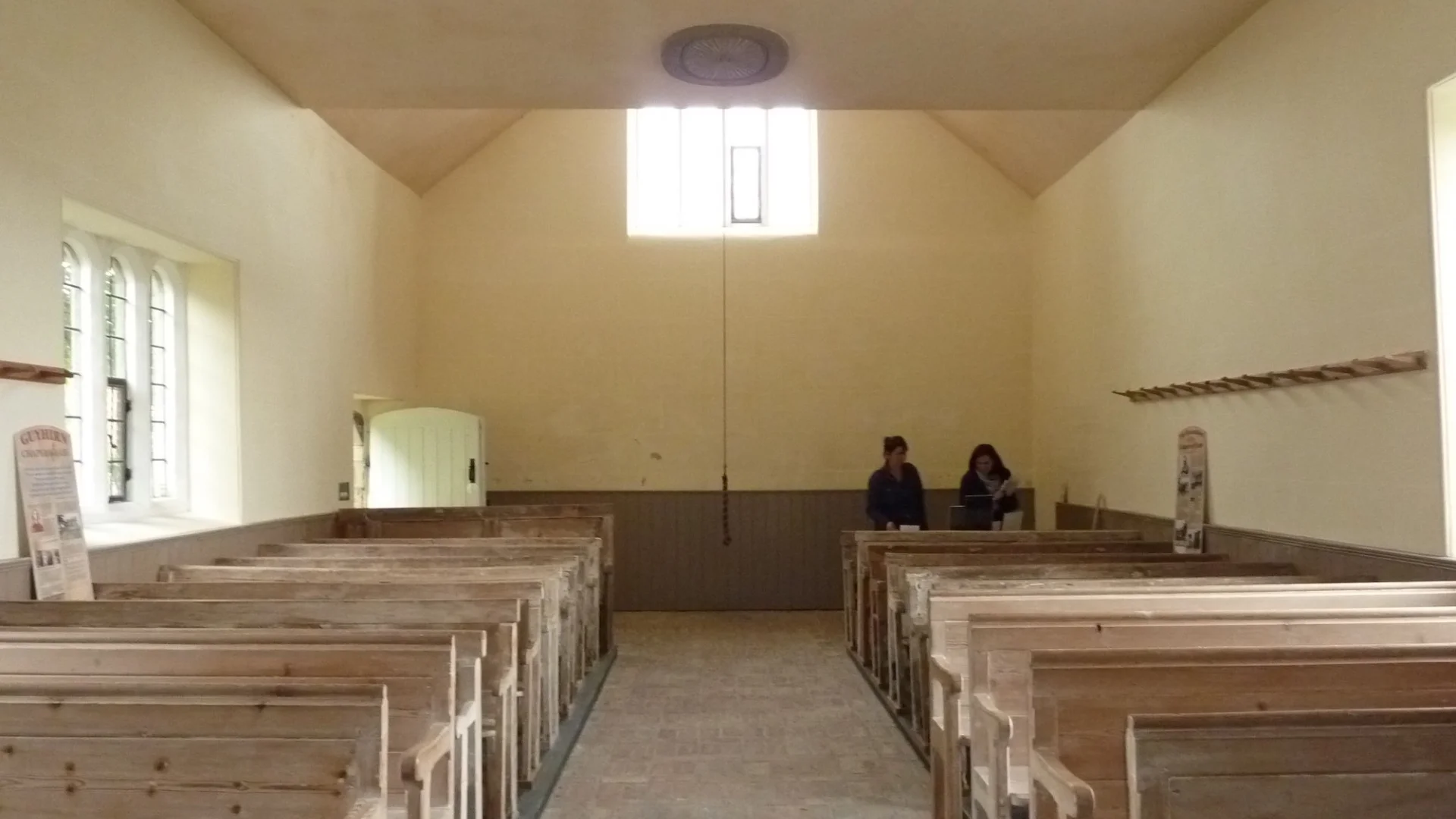 brick and stone nave with plain glass windows and original narrow pews. Two people sit at the back