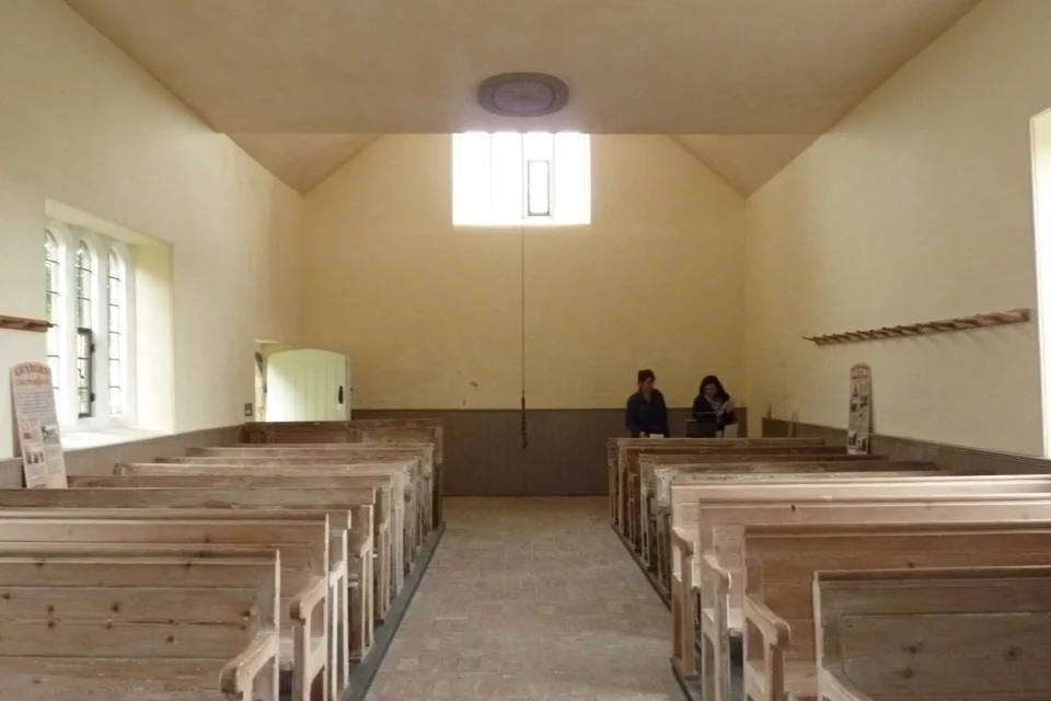brick and stone nave with plain glass windows and original narrow pews. Two people sit at the back