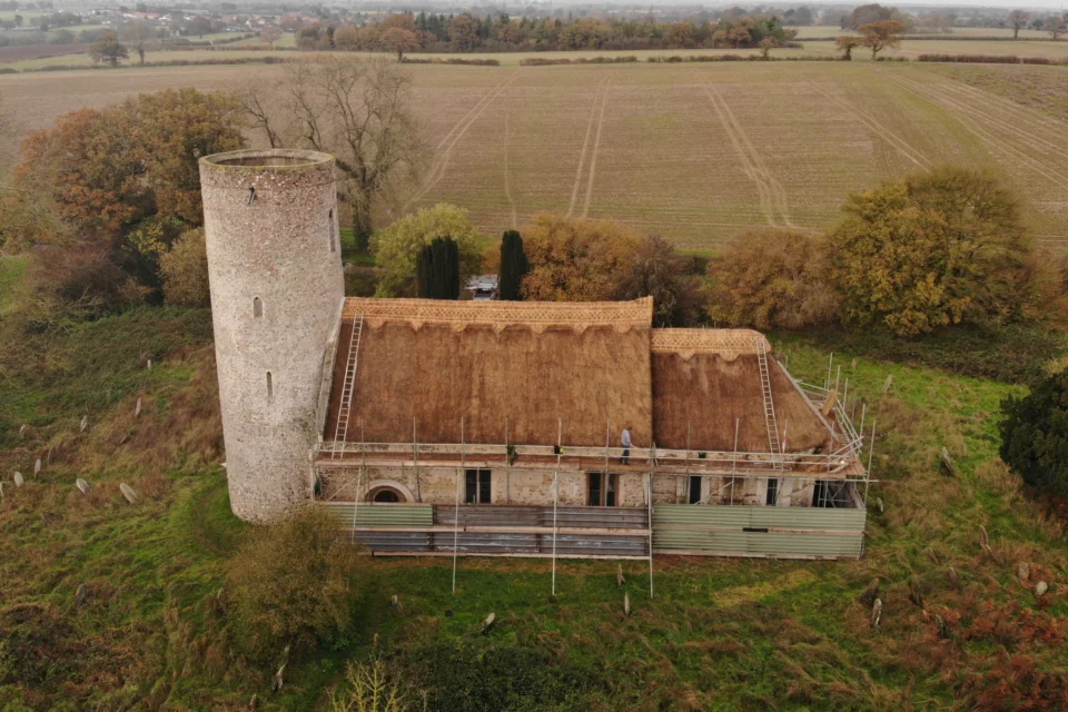A birds eye view of a church roof being repaired and rethatched. Scaffolding surrounds the church and workmen are just visible in the distance.