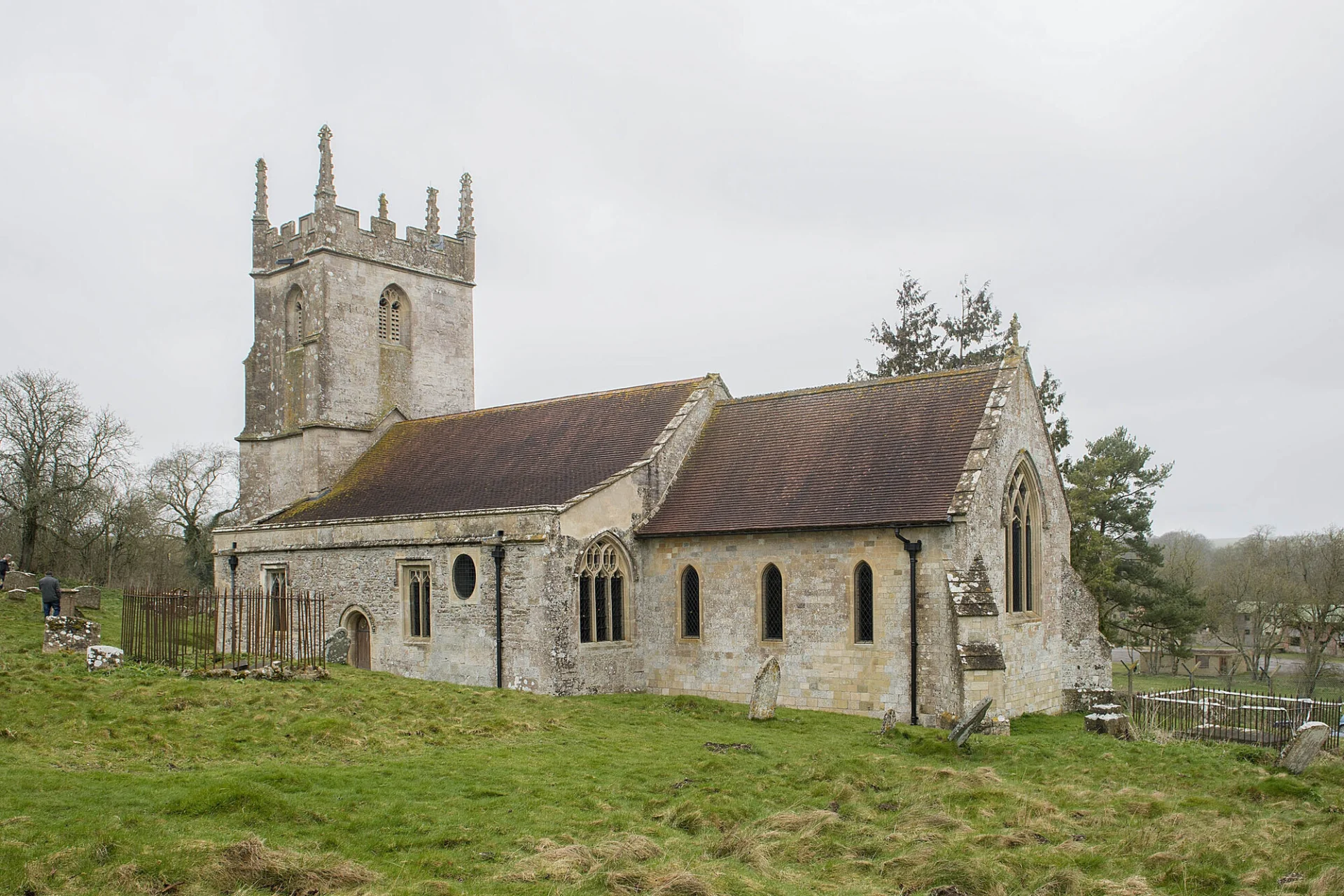 A late-medieval stone church with a five-pinnacled tower stands in the middle of a field below a misty grey sky.