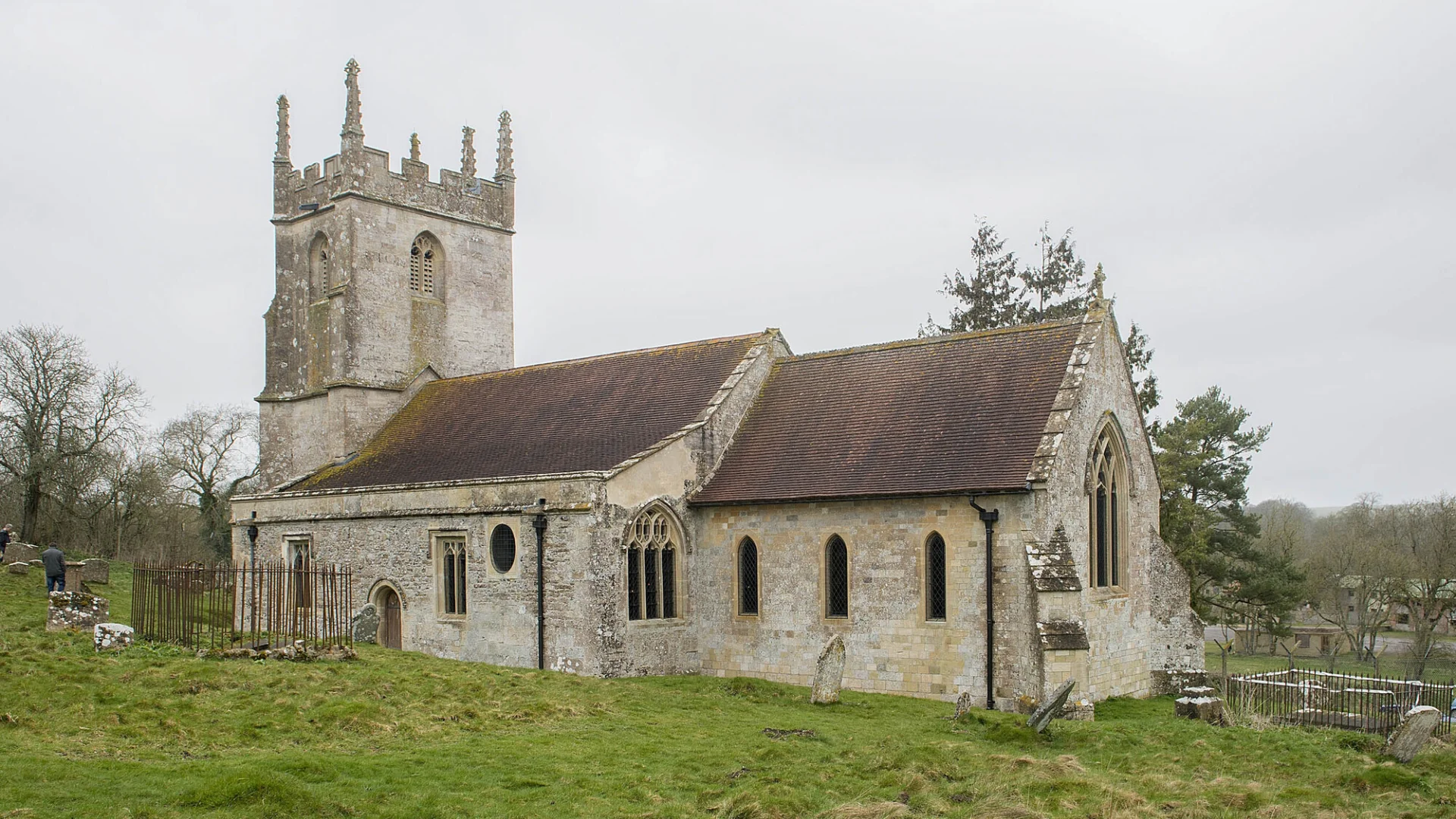 A late-medieval stone church with a five-pinnacled tower stands in the middle of a field below a misty grey sky.