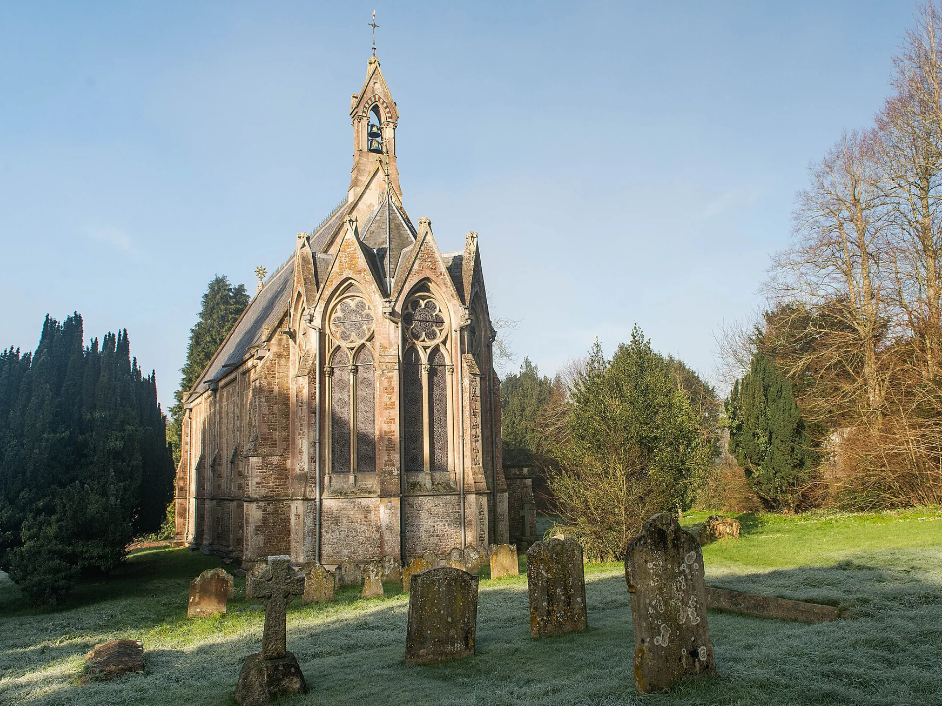 The rear exterior of St Mary's Church on a bright winter's day. The church is surrounded by trees and gravestones, which protrude above the frost-covered grass.