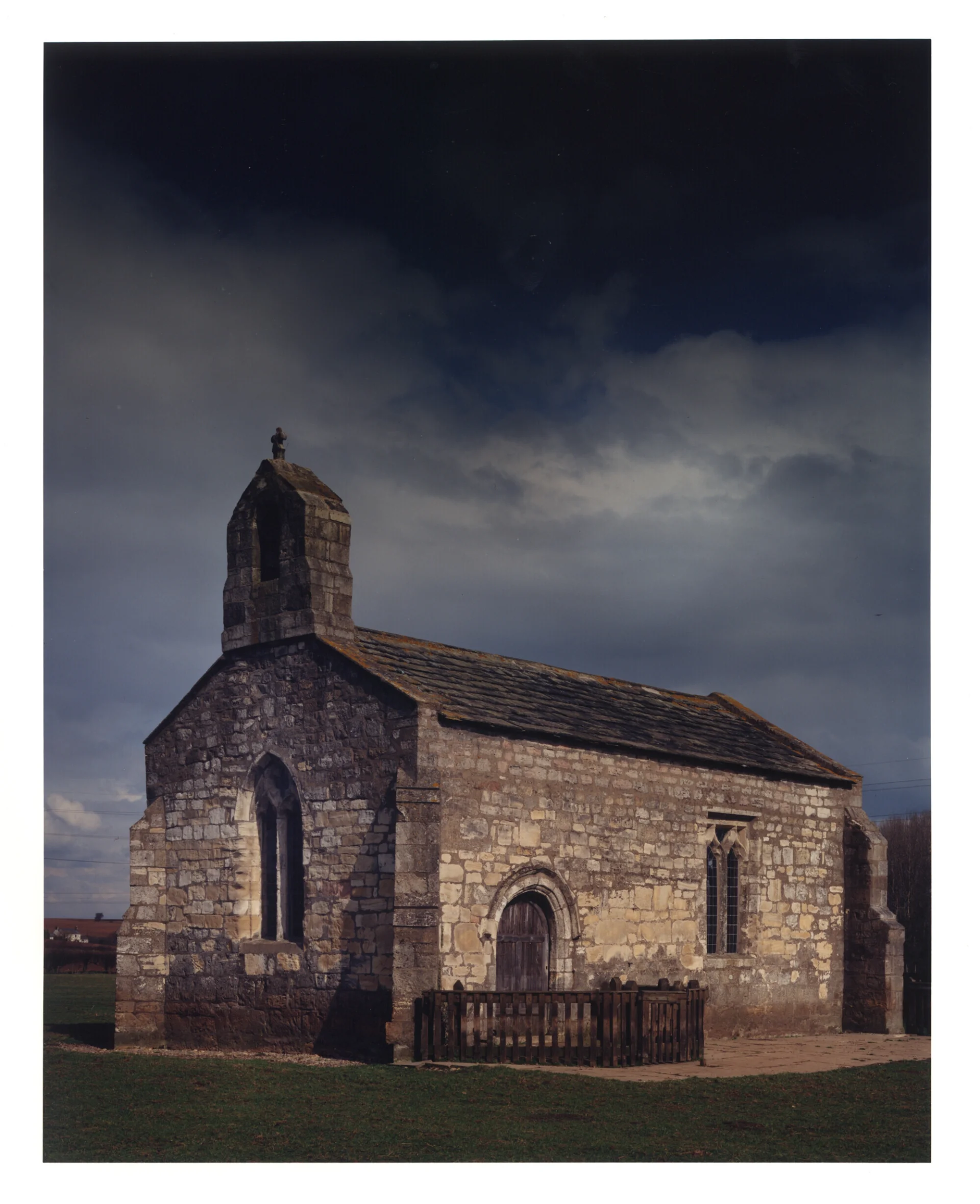 The exterior of a small, rectangular medieval stone chapel in a green field beneath a stormy sky.