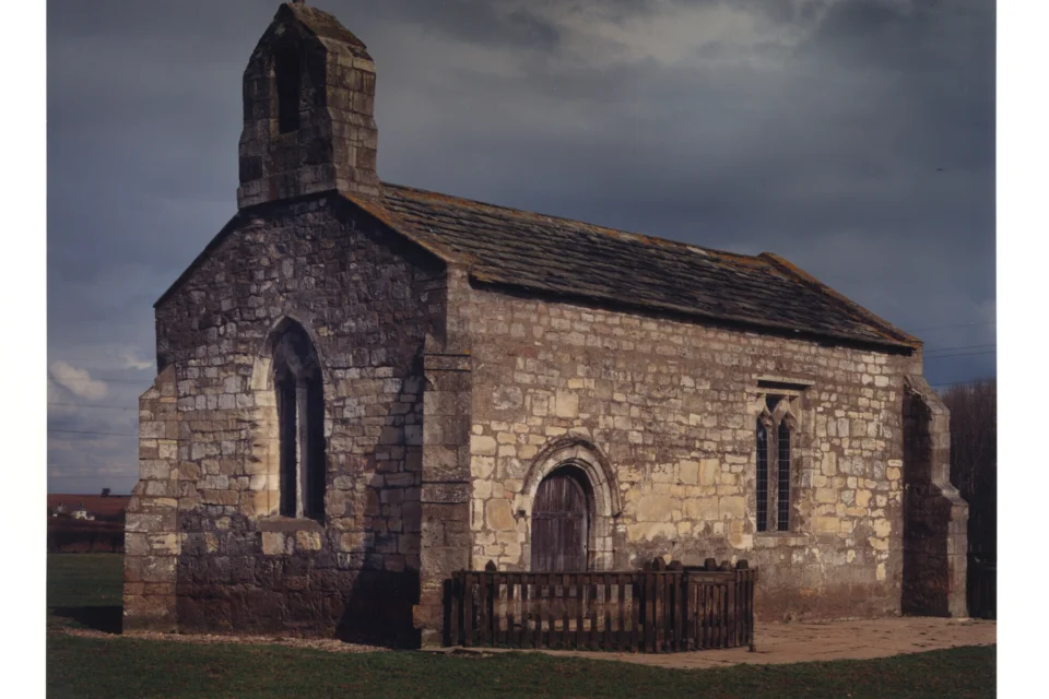 The exterior of a small, rectangular medieval stone chapel in a green field beneath a stormy sky.