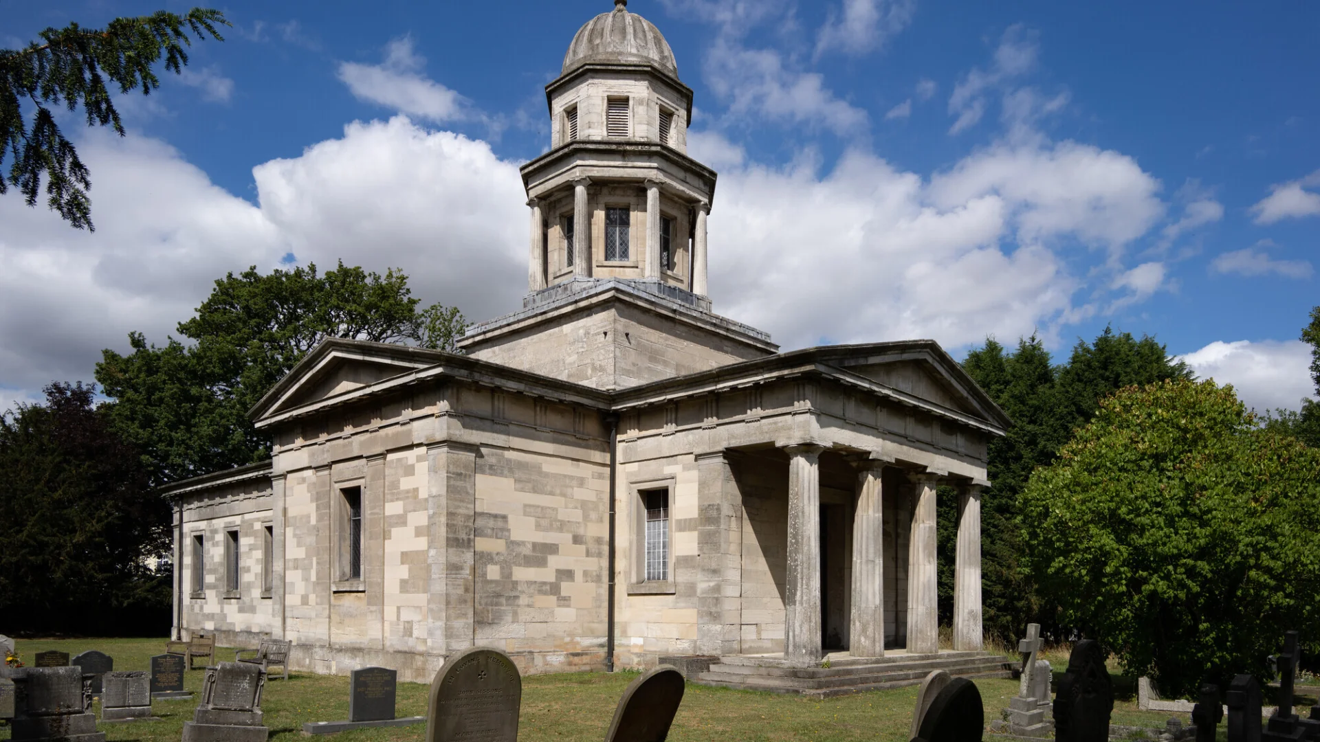A Blue sky shines above a simple stone building, with beautiful cupola on the top and a graveyard to front of the photo.