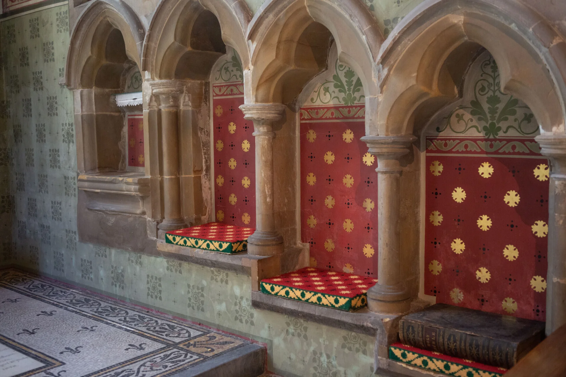 Sedilia with red painted background, with gold reoccurring cross shapes alongside black cross pattern. With colourful embodied pillow in each niche.