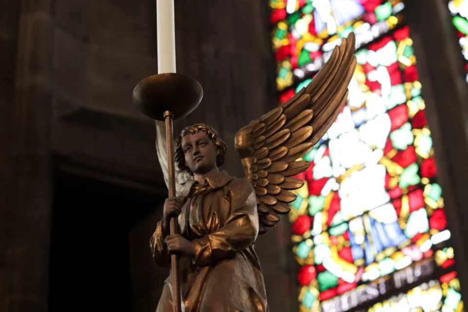 angel statue (possibly brass) holding a candle in front of multi-coloured stained glass window