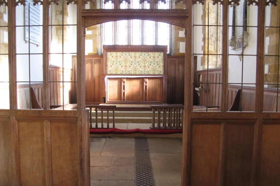 Pews on either side, small gate to alter with decorative tiles in front of a large clear-glass window. Wall monuments on either side of altar.