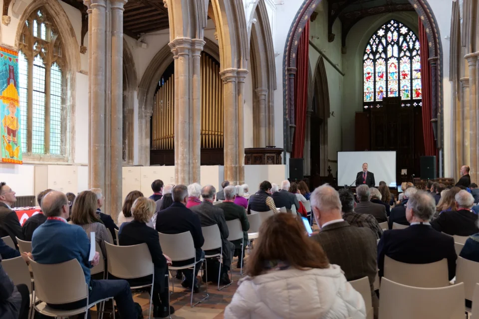 large group of people sit on chairs in the nave watching a presenter with a large screen near the altar. The dazzling stain glass is behind the presenter.