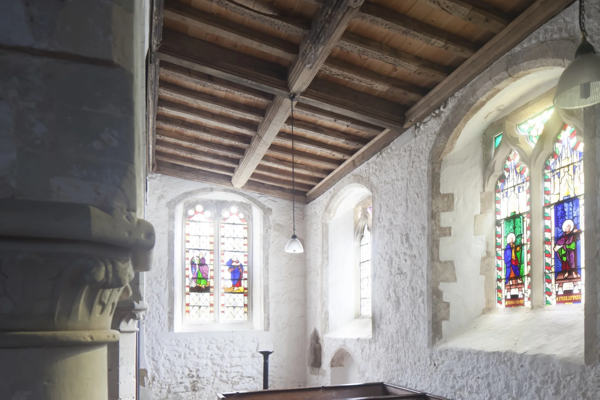 Aisle with white washed walls, windows with portion of stained glass at centre, and slopped timber ceiling