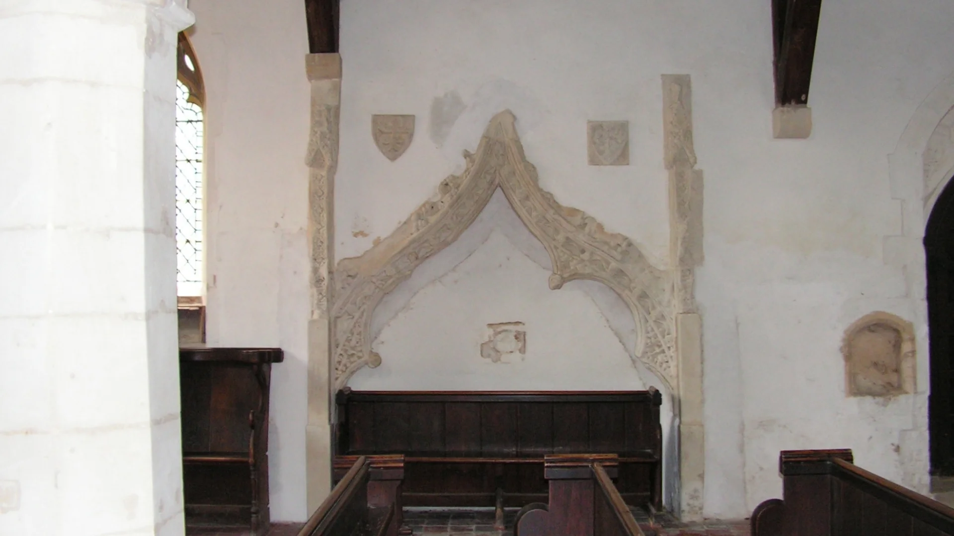 Decorative tomb recess with a pointed arch shape, appearing cream on a white painted wall with two shields in same material on either side of the arch.