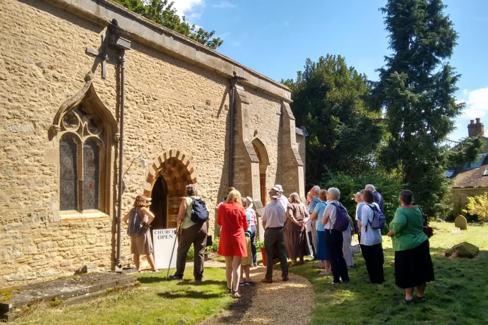Group of people stand in front of the church exterior in the sun, facing the door.