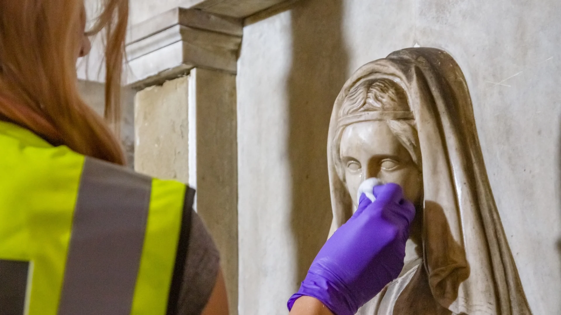 Person in high visibility jacket, cleaning the face of an intricate marble statue of Mary