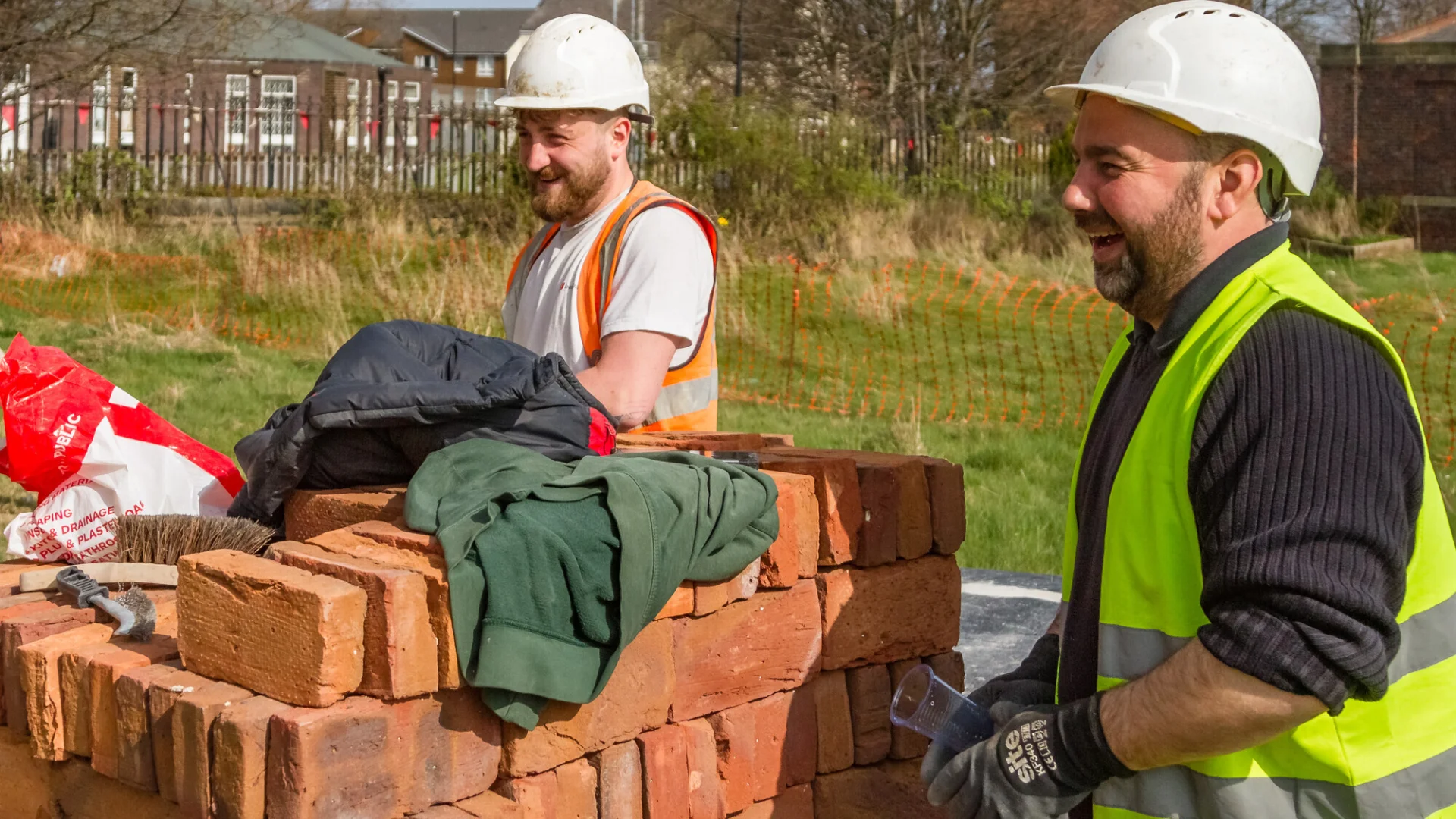 two builders in hard hats and high visibility jacket laughing standing amongst new bricks.