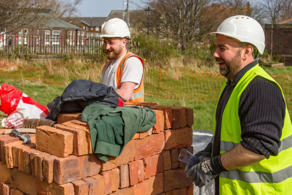 two builders in hard hats and high visibility jacket laughing standing amongst new bricks.