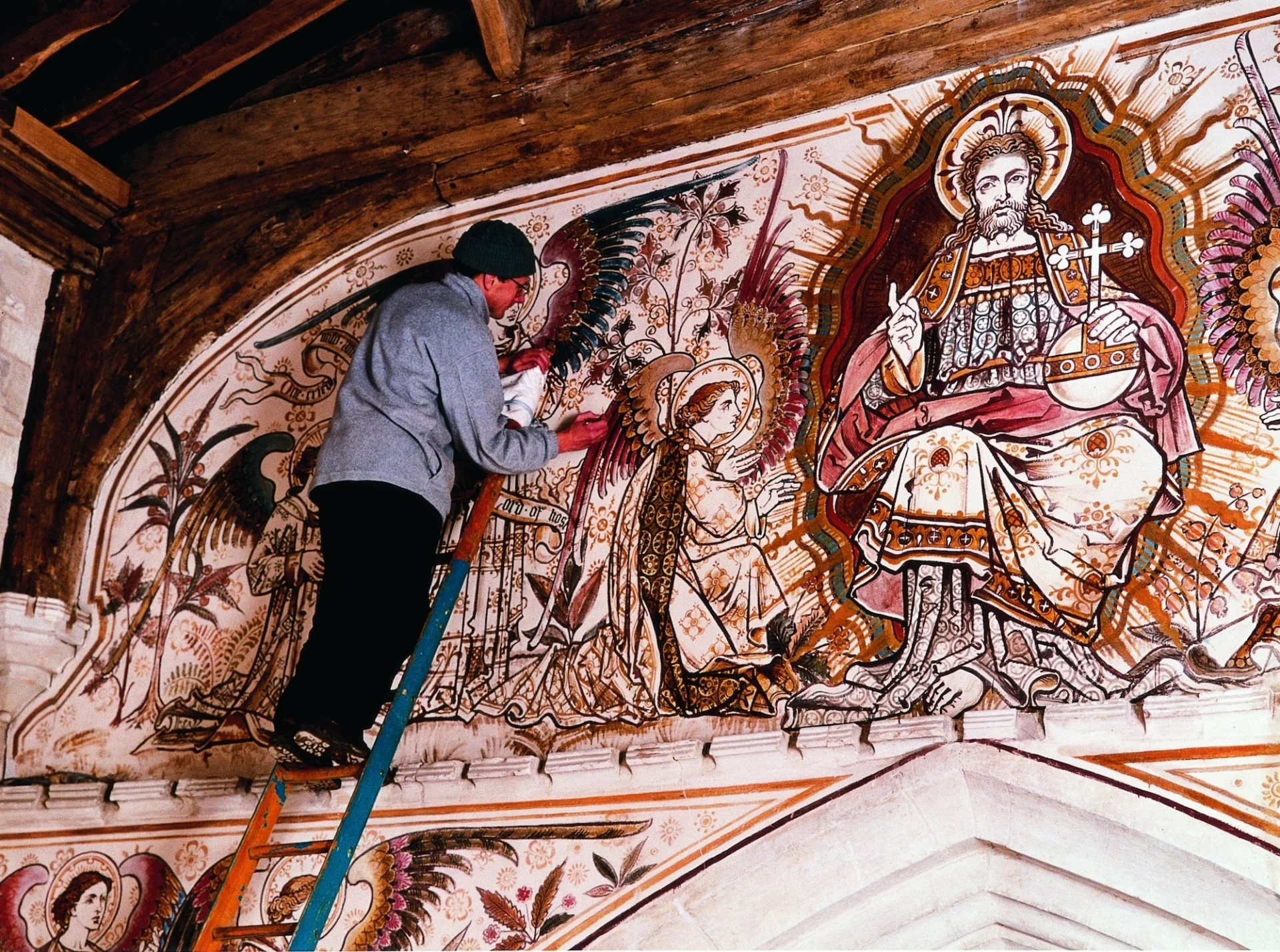 man on ladder is restoring vibrant red and gold wall paintings with Jesus (as King of heaven) at centre and an angel kneeling to him.