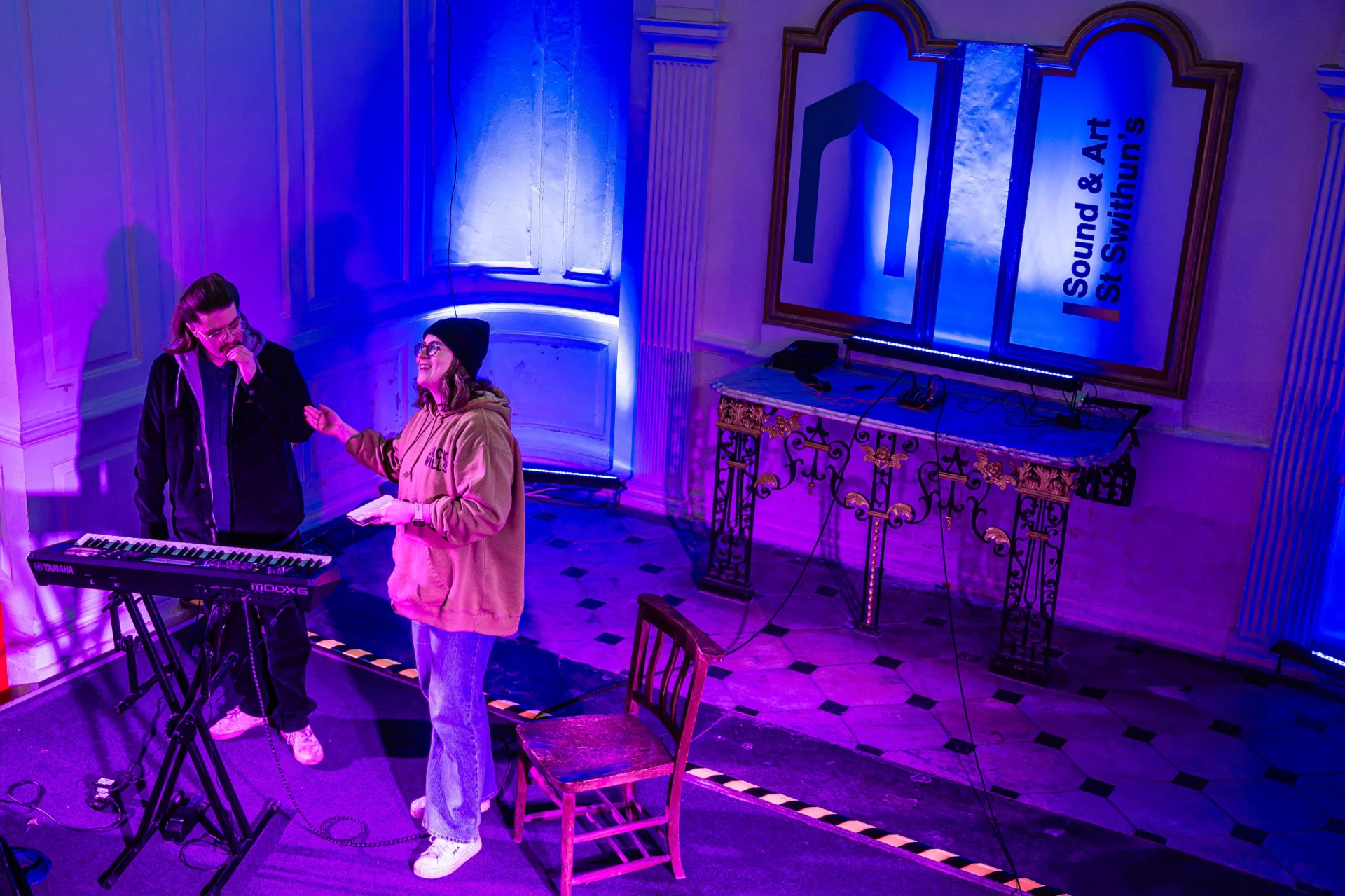 two performers standing, rehearsing on the stage. One singing with sheet music in hand and the other listening next to their keyboard. There is a small table at the back with beautiful metal legs and two wooden signs above with the St Swithun's logo and black text, 'Sound and Art St Swithun's'.