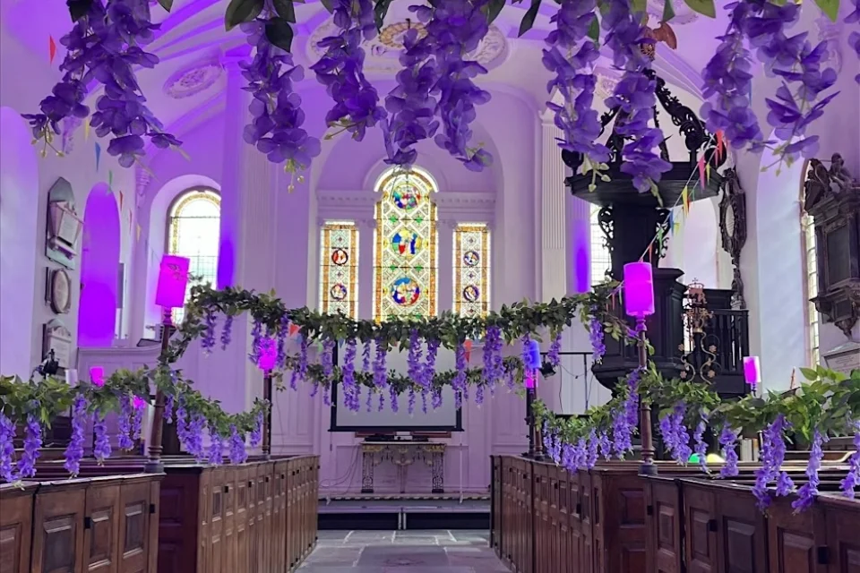 Large nave draped with fake lilac flowers like bunting across the aisle and lit with a lilac light. The box pews and stone floor as well as large stained glass window behind the altar are also clear.