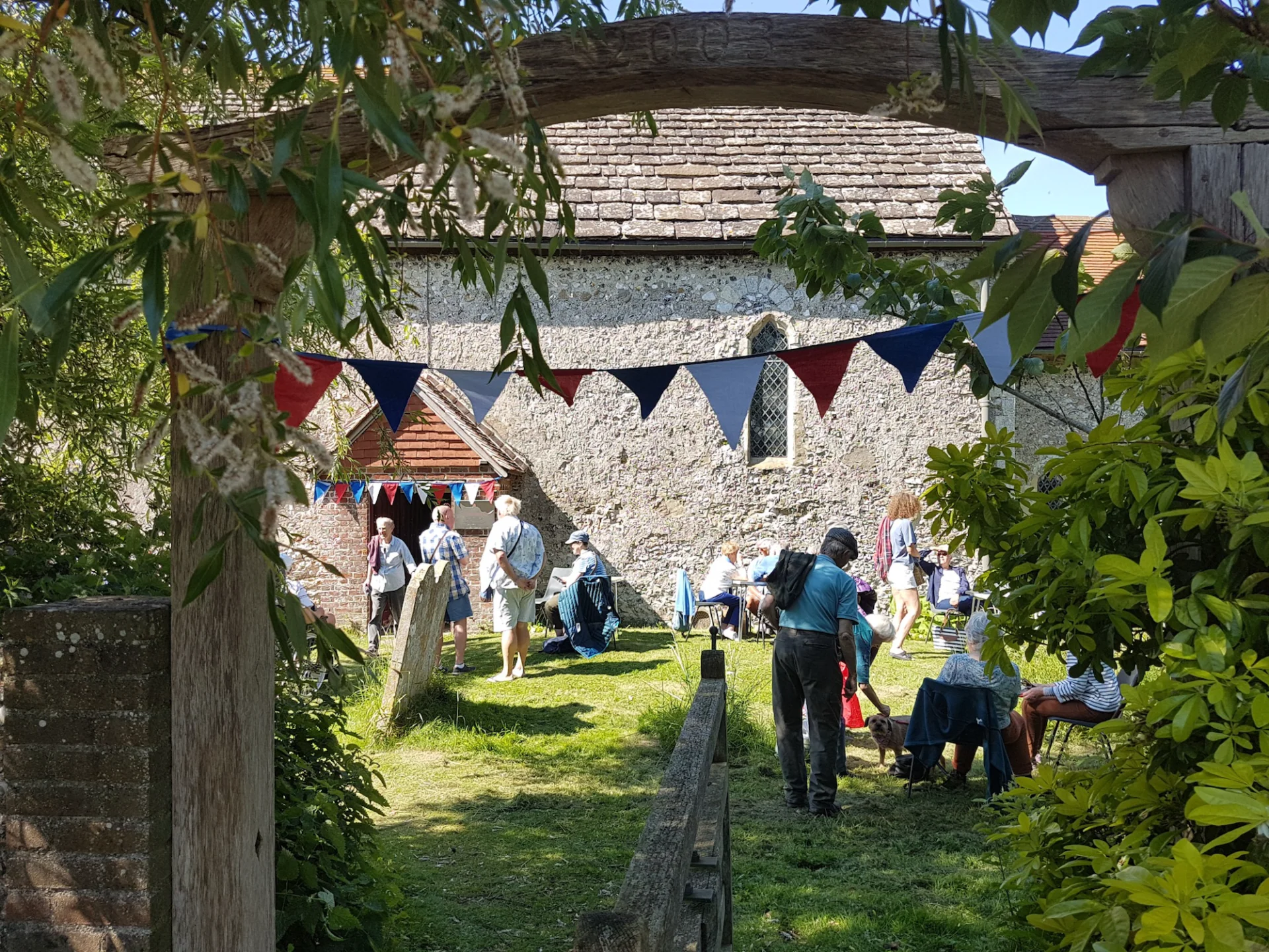 glimpse of people enjoying a garden party beside a church, through a wooden archway entrance. There is colourful bunting and chairs.