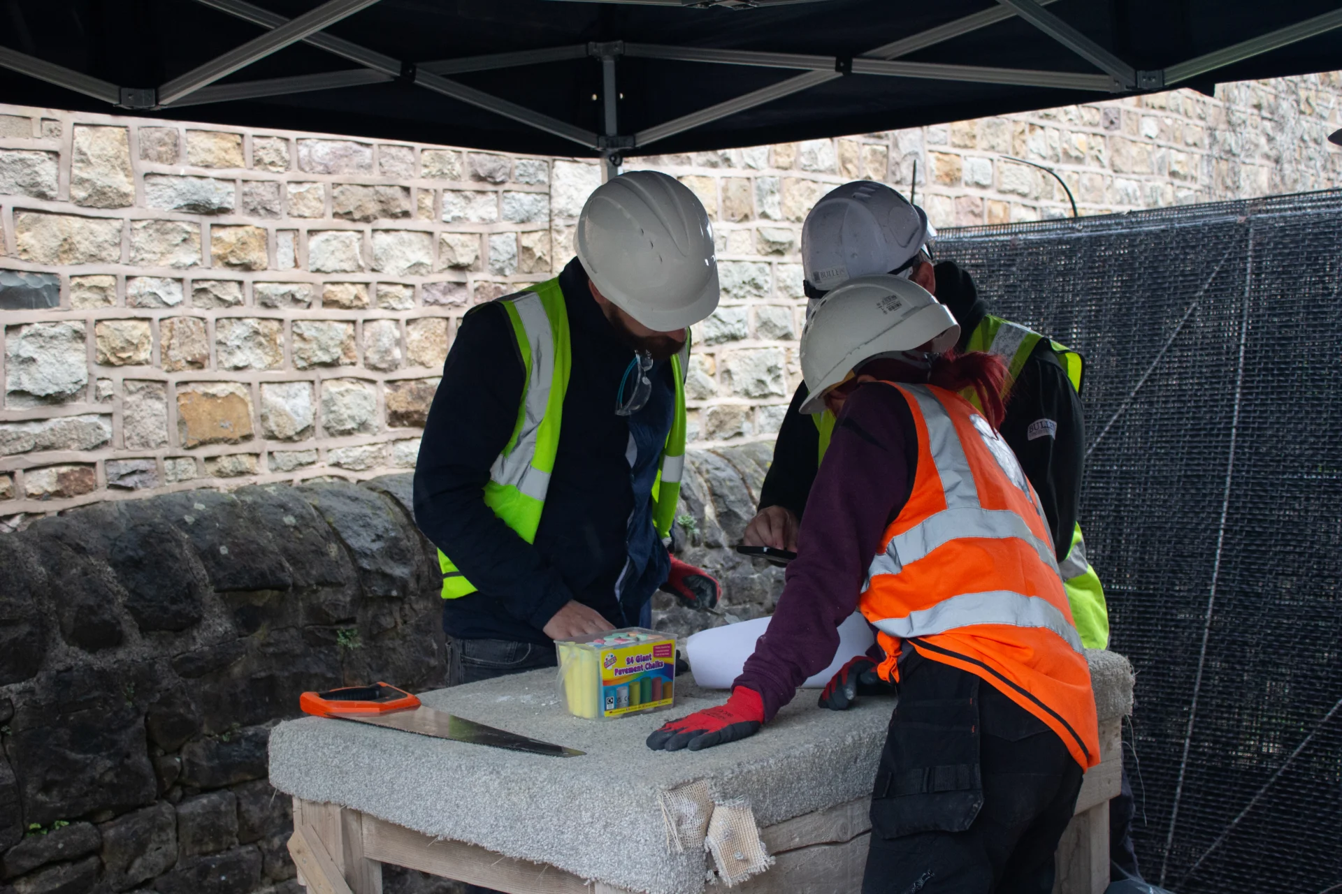 three people in high visibility jackets and hard hats crowd around a table outside, looking  at plans