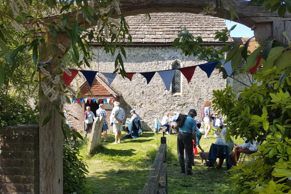 glimpse of people enjoying a garden party beside a church, through a wooden archway entrance. There is colourful bunting and chairs.