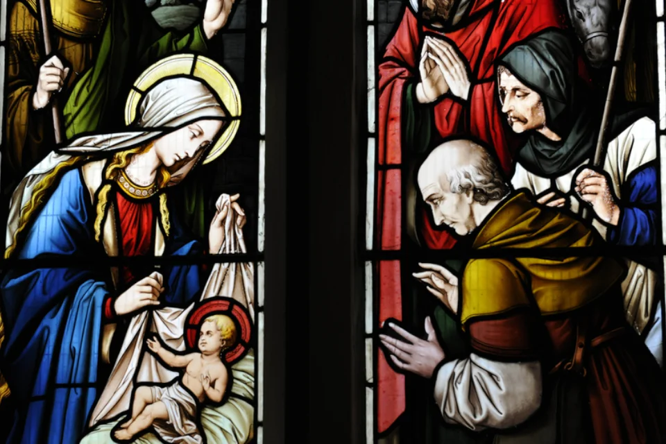 brightly coloured traditional stained glass with Mary bent over the baby Jesus preparing to swaddle him and the wise men on the right hand side.