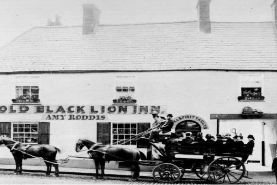 black and white old image of the Old Black Lion Inn with the name Amy Roddis on the front, and a cart with two horses and rides with top and bowler hats