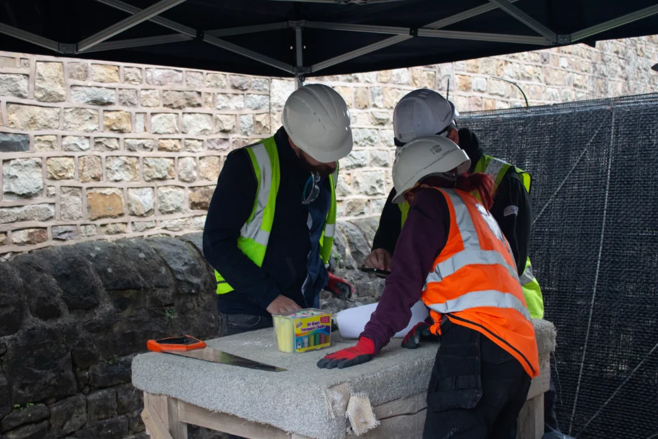 three people in high visibility jackets and hard hats crowd around a table outside, looking  at plans